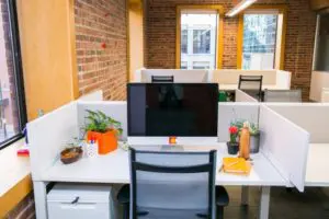 Modern office cubicle with a computer monitor, chair, and desk holding potted plants, a bowl, pen holder, notebook, and water bottle; surrounded by white dividers, with additional cubicles and large windows providing natural light and a view of a nearby building.