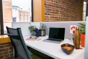 Modern office workspace with a desk featuring a laptop, notebook, pen, bowl of snacks, water bottle, and two small potted plants; a chair is placed in front of the desk, which sits beside a window overlooking a brick building, with a matching brick wall behind.